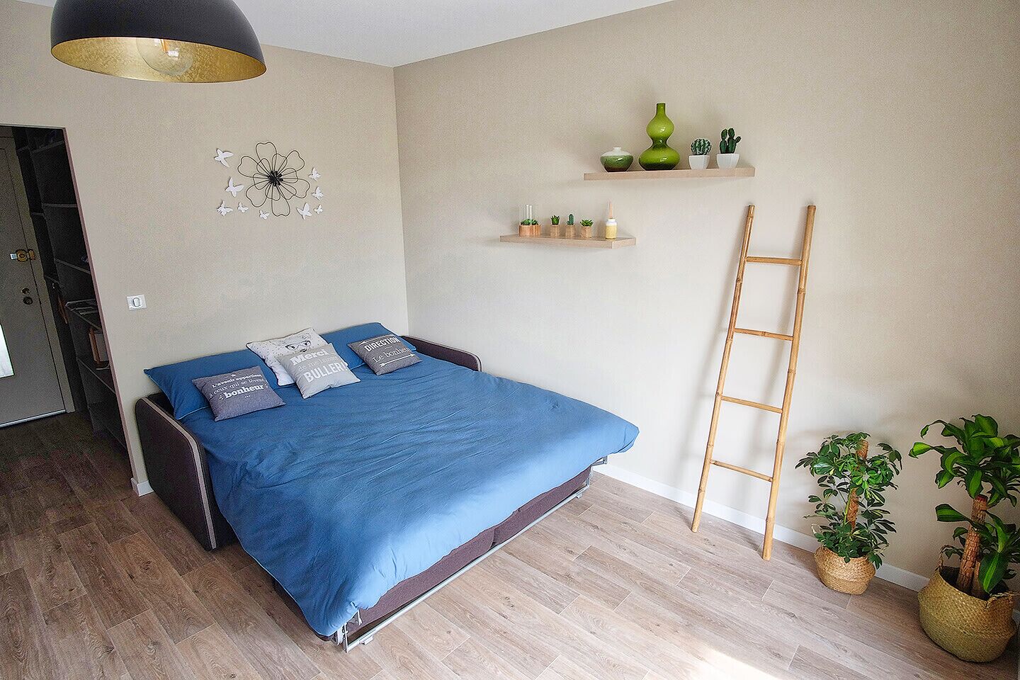 Un dormitorio con cama, almohadas, estante en la pared con plantas, una escalera y una puerta a otra habitación.