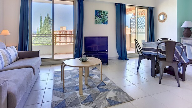 A modern living room with a sofa, a TV, a dining table with chairs, and a balcony with blue curtains. - (029) L'entre deux à 4 (Antibes)