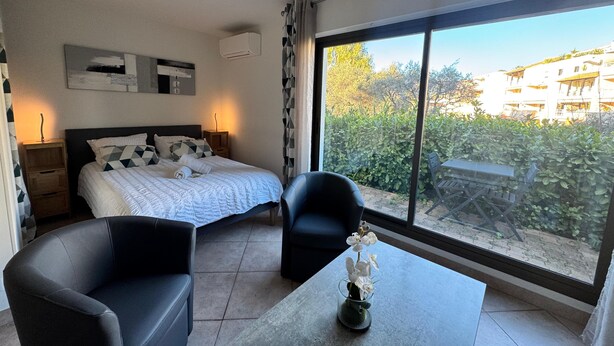 A modern bedroom with a large window, a bed with geometric-patterned pillows, a side table, a chair, and a vase with flowers.