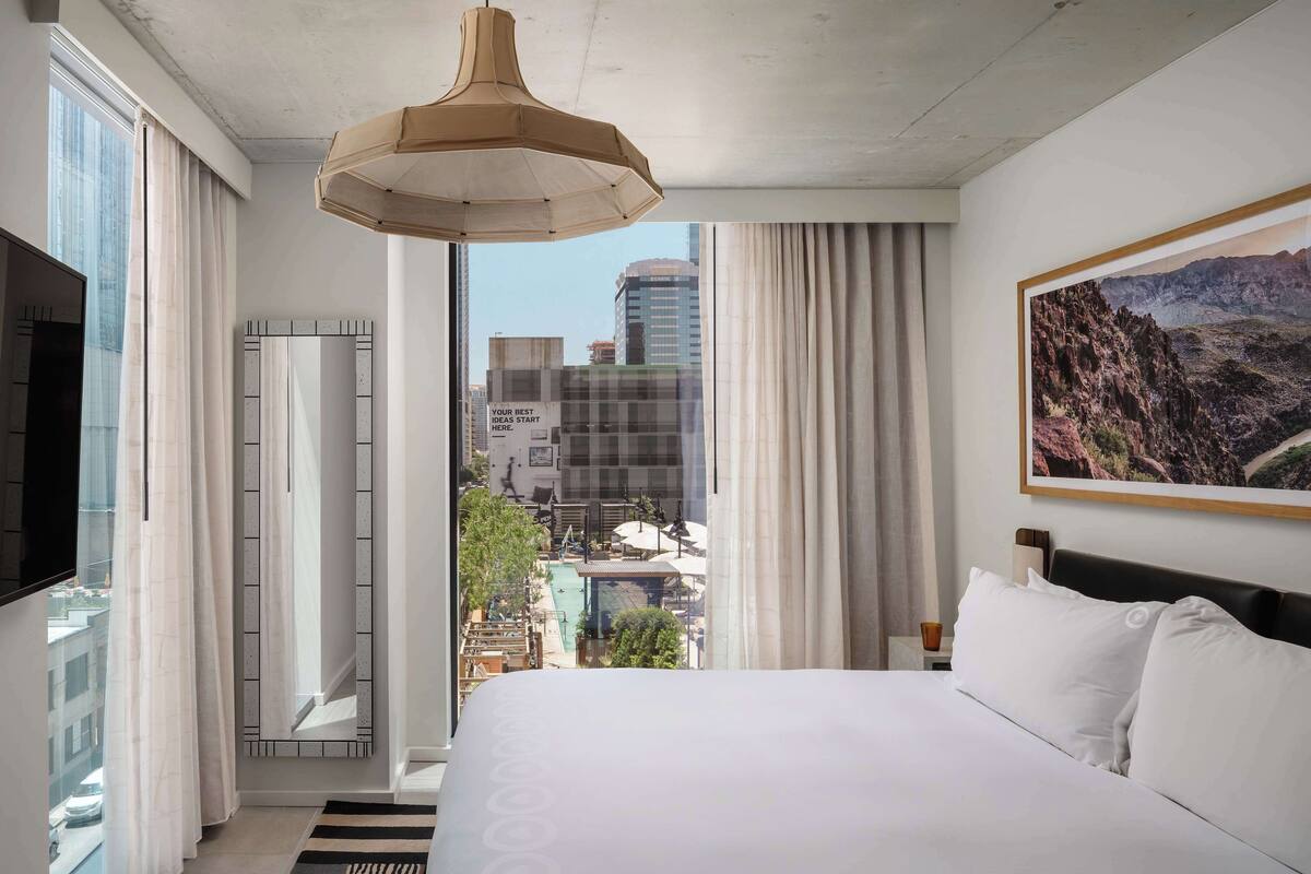 Une chambre d’hôtel avec un grand lit, un téléviseur fixé au mur, une vue sur la ville et un tableau accroché au mur.