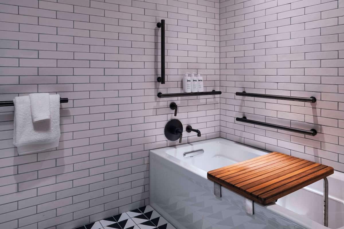 Une salle de bain avec des murs en briques blanches, un sol carrelé en noir et blanc, un banc en bois, une baignoire et un porte-serviettes.