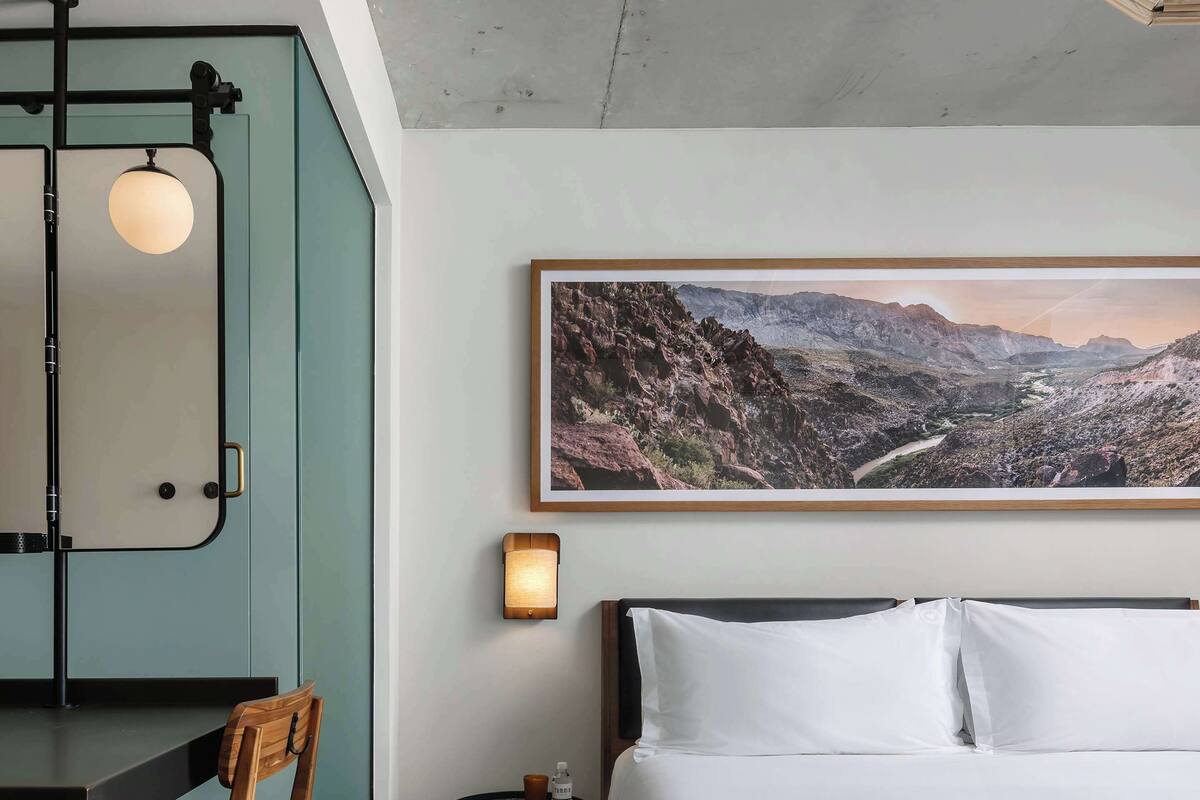 Une chambre d’hôtel avec un lit, un bureau, une chaise, une petite table avec une lampe et un grand tableau représentant un paysage accroché au mur.