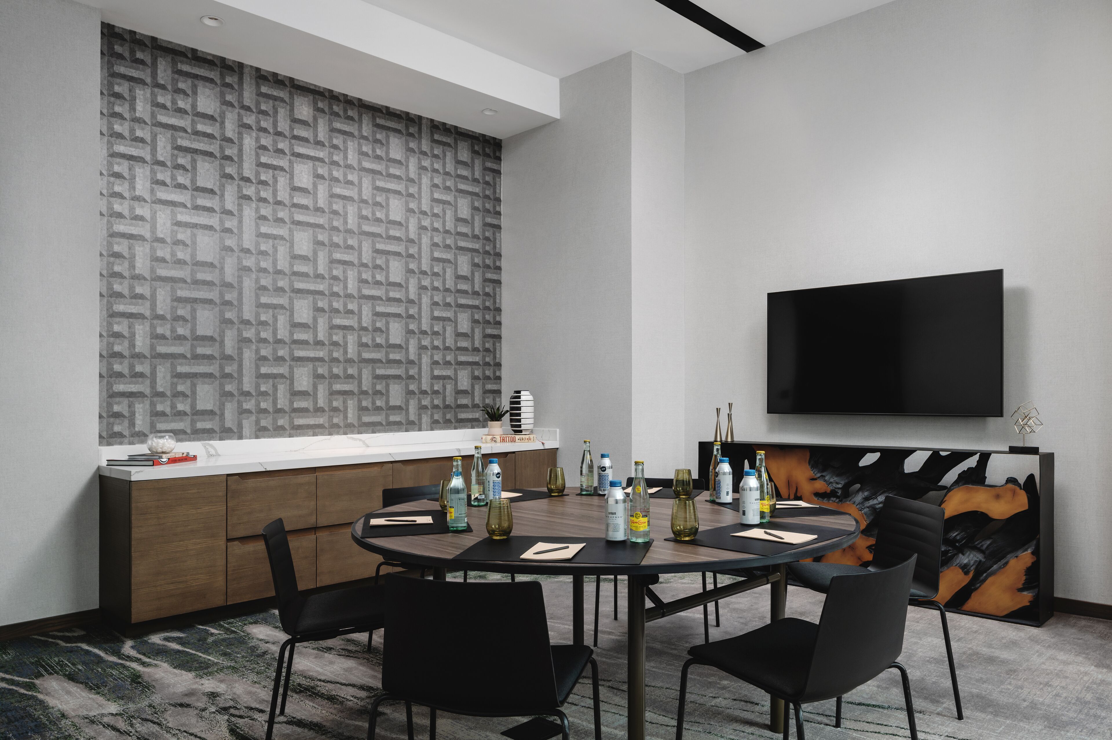 A modern conference room with a round table, black chairs, a large TV, and a patterned wall.