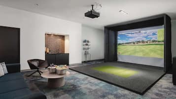 A modern living room with a large screen displaying a golf course, a comfortable sofa, a coffee table with snacks, and a projector ceiling light.