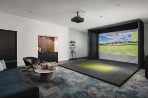 A modern living room with a large screen displaying a golf course, a comfortable sofa, a coffee table with snacks, and a projector ceiling light.