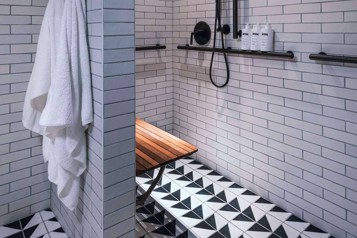 Une salle de bain moderne avec des murs en briques blanches, un sol carrelé en noir et blanc, un porte-serviettes, un banc en bois et une douche équipée d’un pommeau de douche à main et de quelques produits placés sur une étagère.