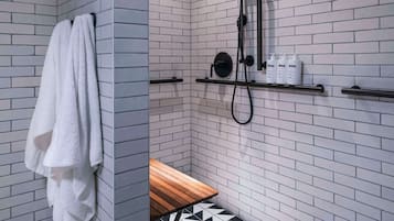 Un baño moderno con paredes de ladrillo blanco, piso de baldosas blanco y negro, un perchero, un banco de madera y una ducha con cabezal de ducha manual y botellas en un estante.