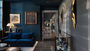 A modern hotel lobby with a blue sofa, a coffee table, and a lamp. There is a hallway leading to another room with a glass railing and decorative items.