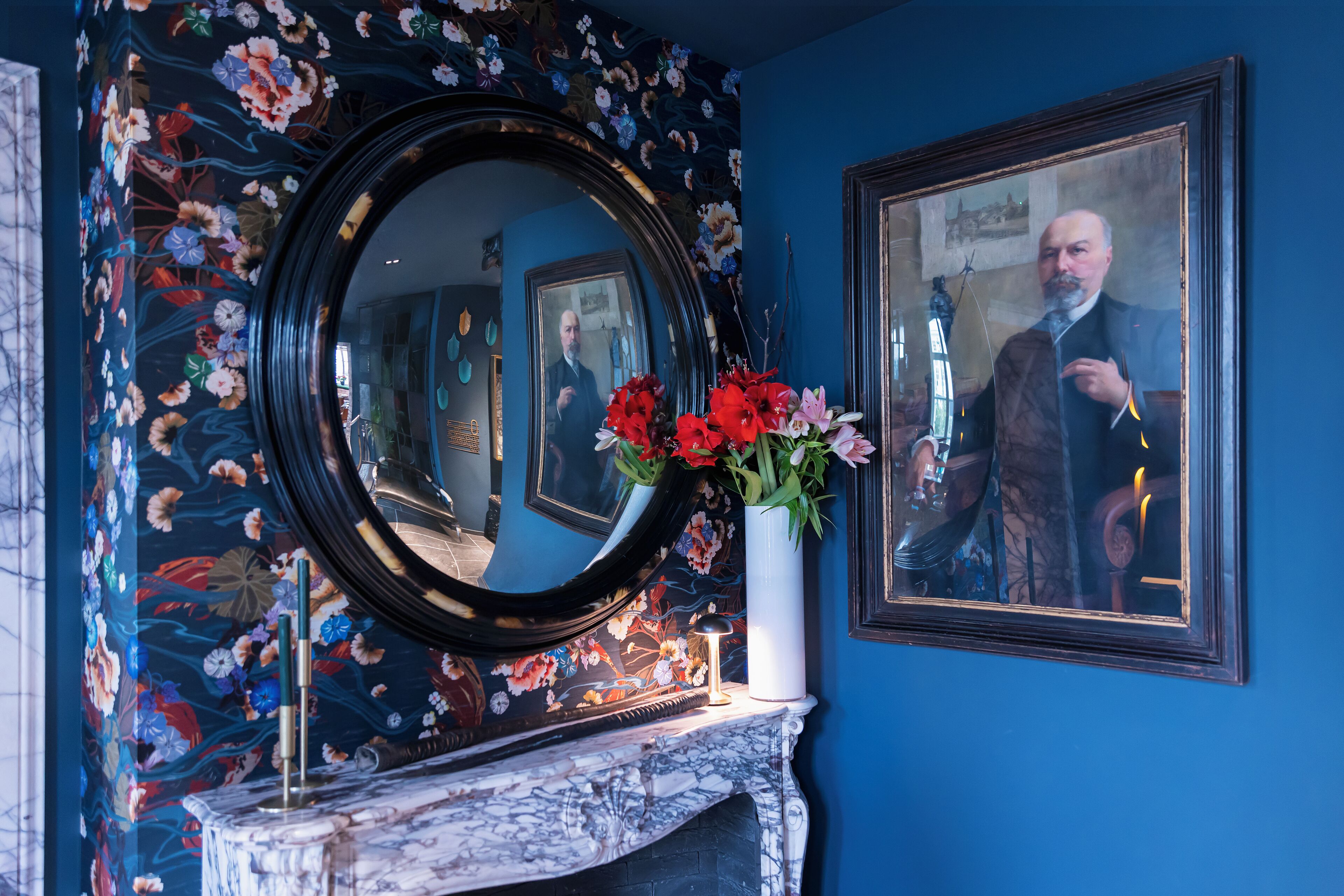 A room with a floral wallpaper, a round mirror, a vase with flowers, and a framed portrait on the wall.