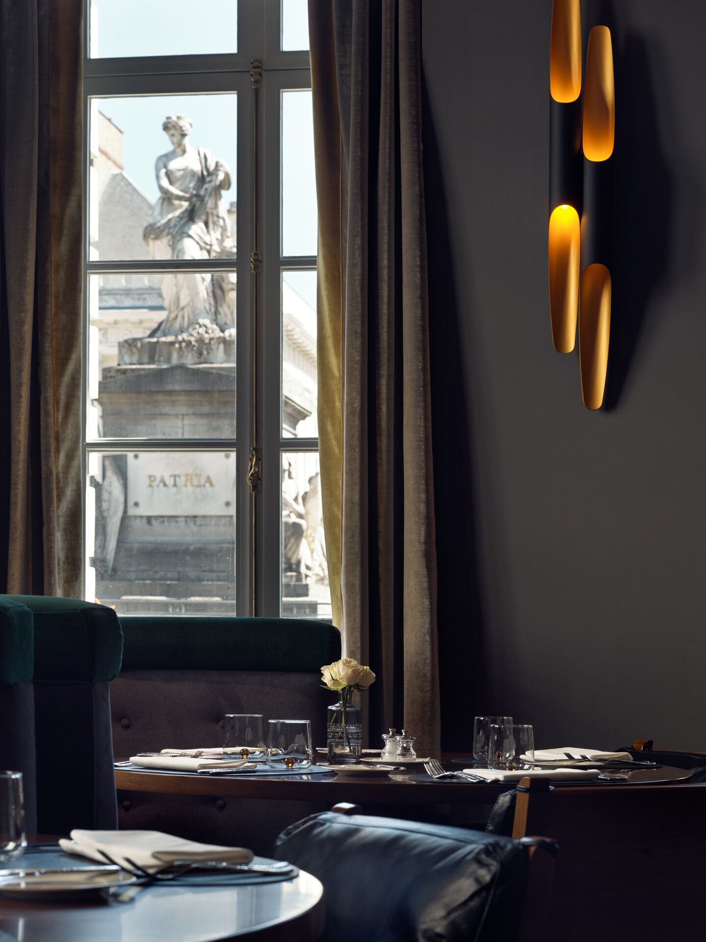 A dining area with a view of a statue through a window, a vase with a single flower, and a table set for two.