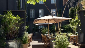 An outdoor patio with wooden tables and chairs, surrounded by potted plants and a large umbrella.