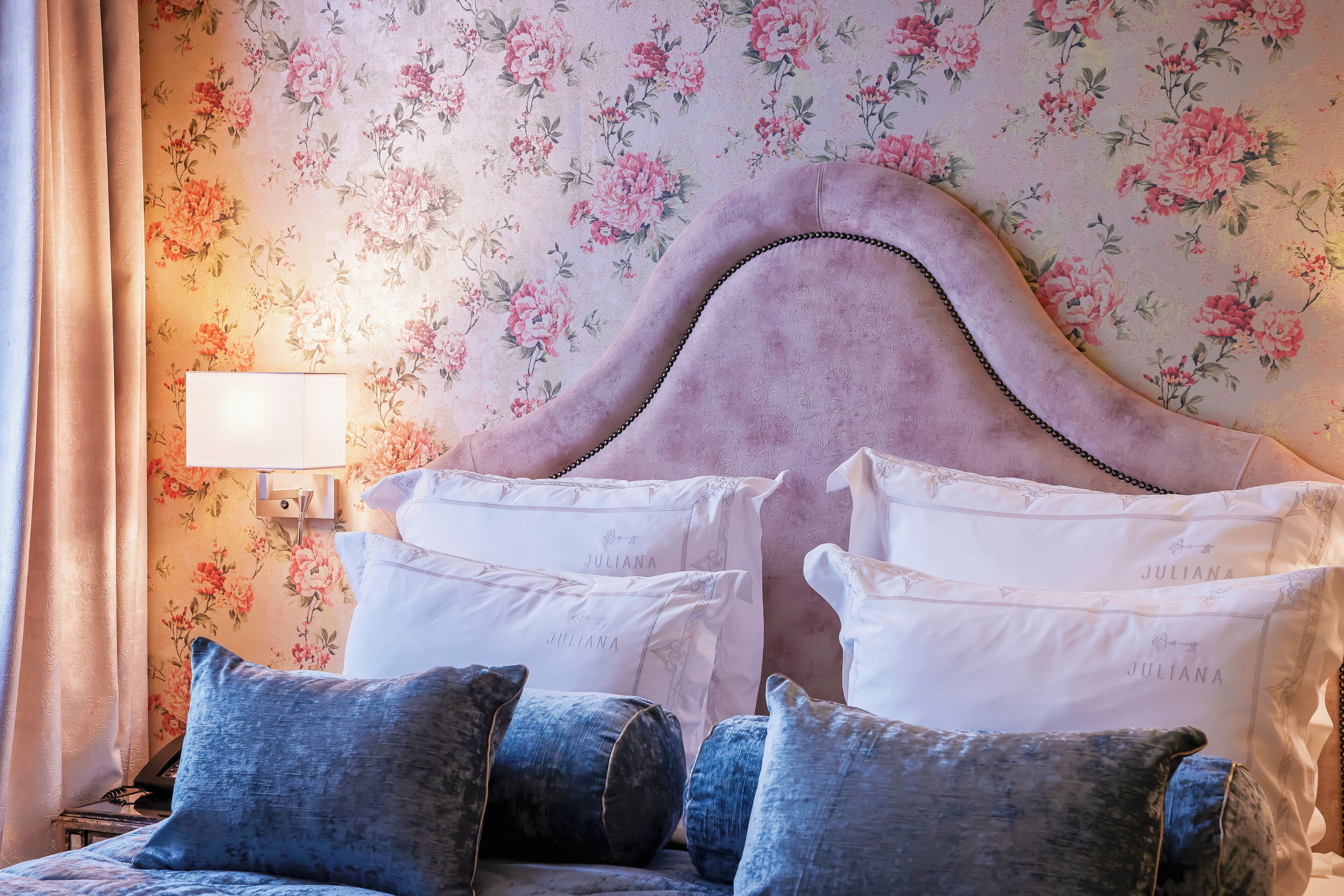 A bedroom with a floral wallpaper, a pink upholstered headboard, and multiple pillows.