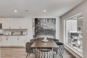 Een moderne keuken met witte kasten, een grote eettafel en een zwart abstract schilderij aan de muur.