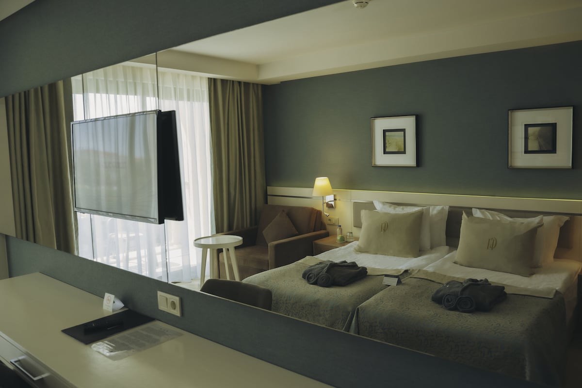 A hotel room with a bed, a desk, a chair, a small table, a lamp, and a window with curtains.