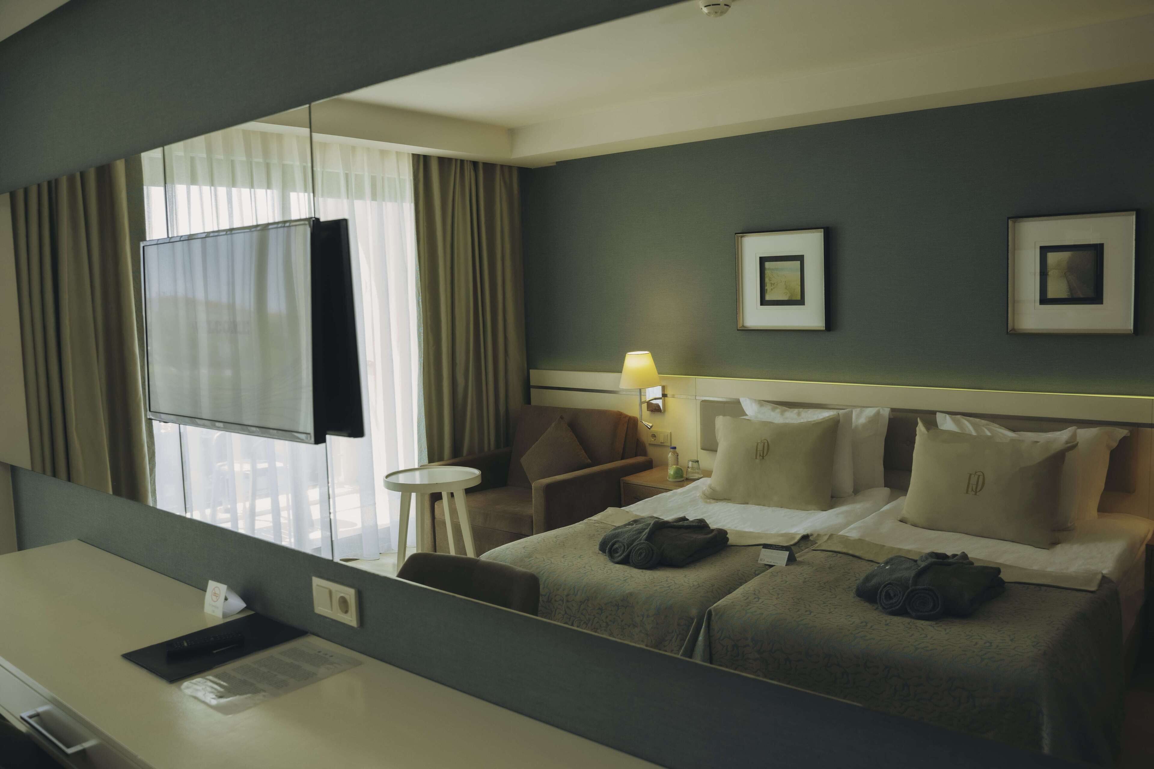 A hotel room with a bed, a desk, a chair, a small table, a lamp, and a window with curtains.