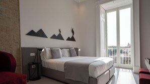 A modern bedroom with a large bed, a red chair, a floor lamp, and a city view through the window. - Ciao Bellini (Naples)