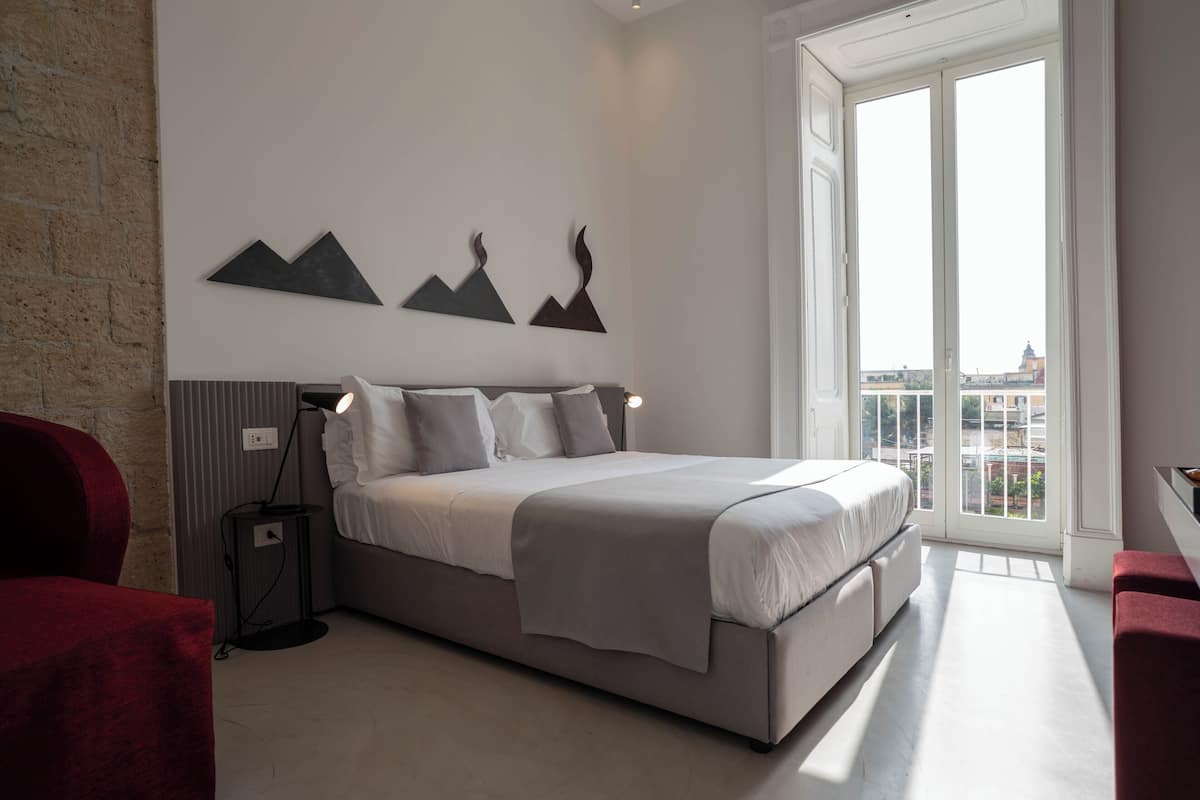 A modern bedroom with a large bed, a red chair, a floor lamp, and a city view through the window.