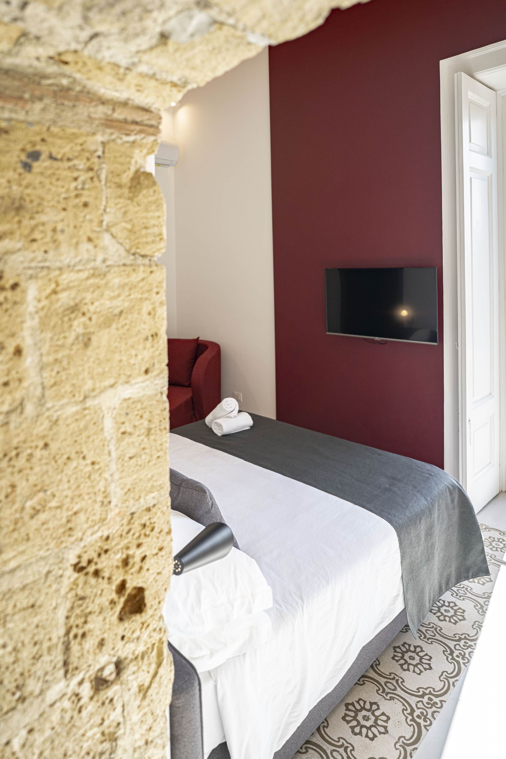 Habitació d'hotel amb paret de pedra, un llit amb mantell blanc i gris, una cadira vermella i un televisor de pantalla plana muntat a la paret.