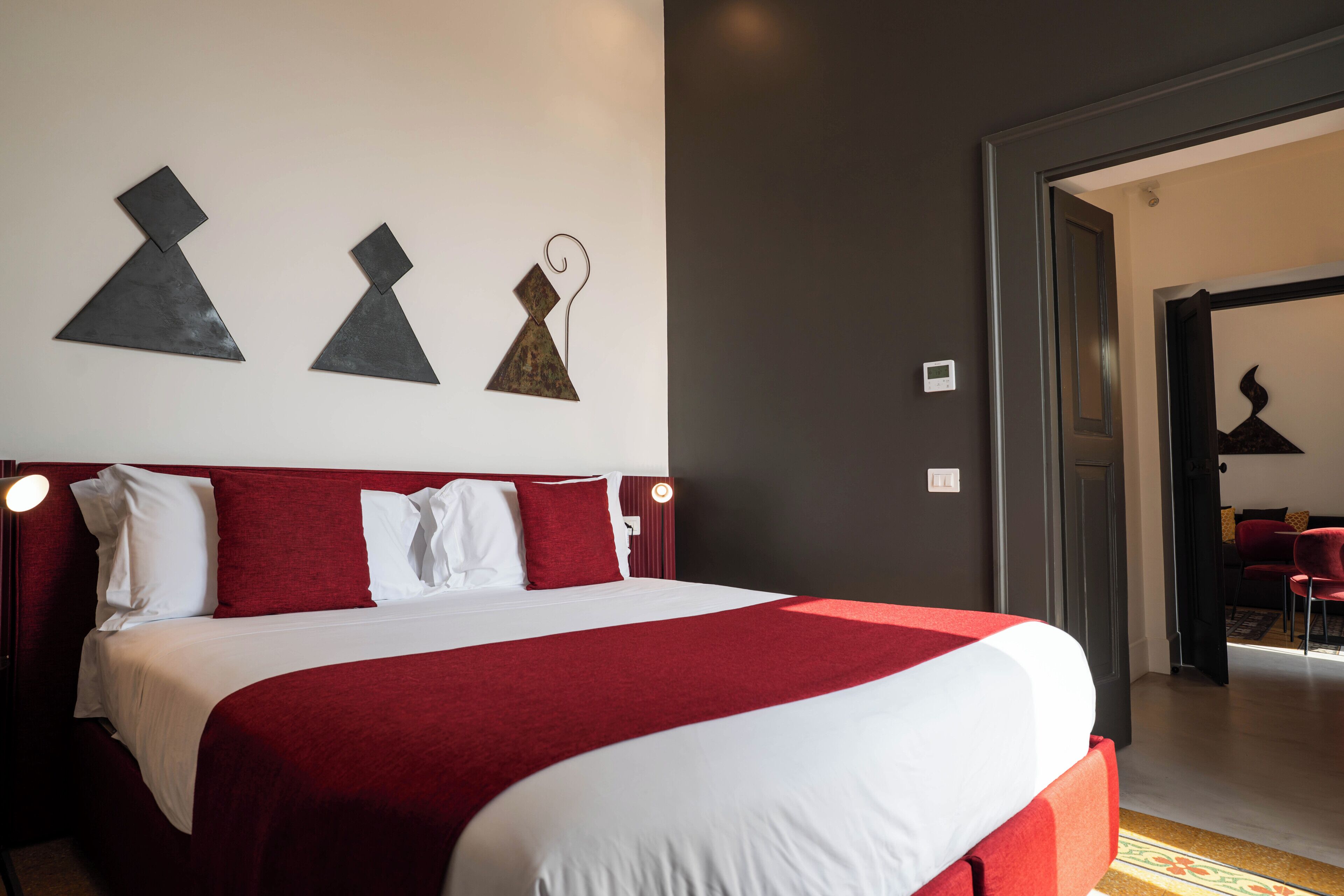 A hotel room with a large bed, red and white bedding, a modern wall art piece, and a view into the adjacent room.