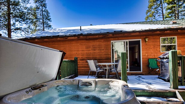 Una cabaña de madera con hidromasaje, mobiliario exterior y un techo cubierto de nieve.