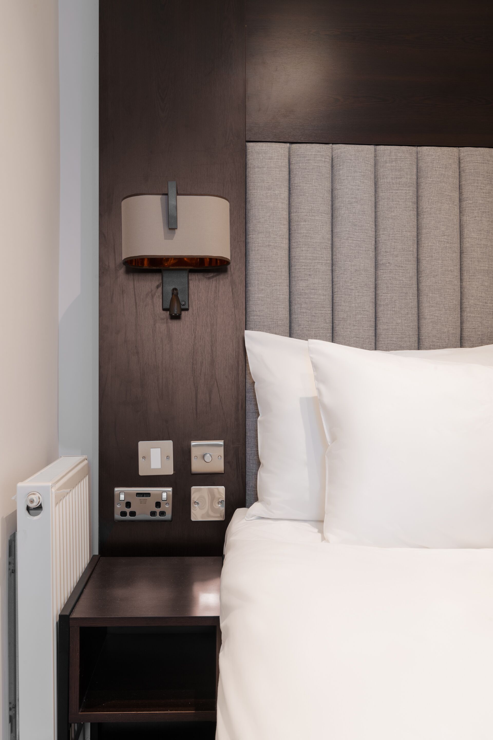 Een hotelkamer met een hoofdeinde van donker hout, een nachtkastje met lamp, en een wit bed.