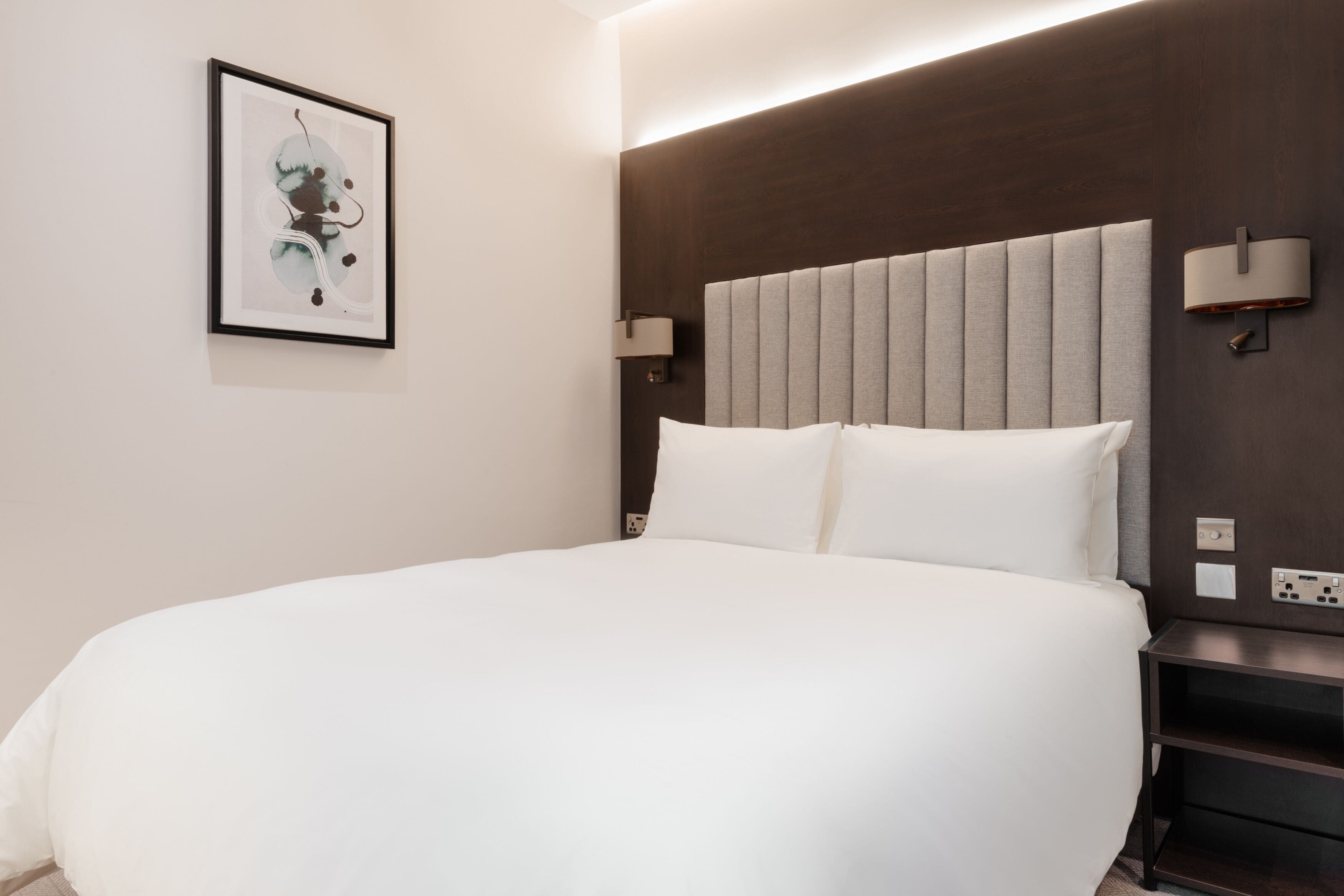 Ein Hotelzimmer mit einem großen Bett, einer Kopfteilbeleuchtung, einem Nachttisch und einer Wandlampe.