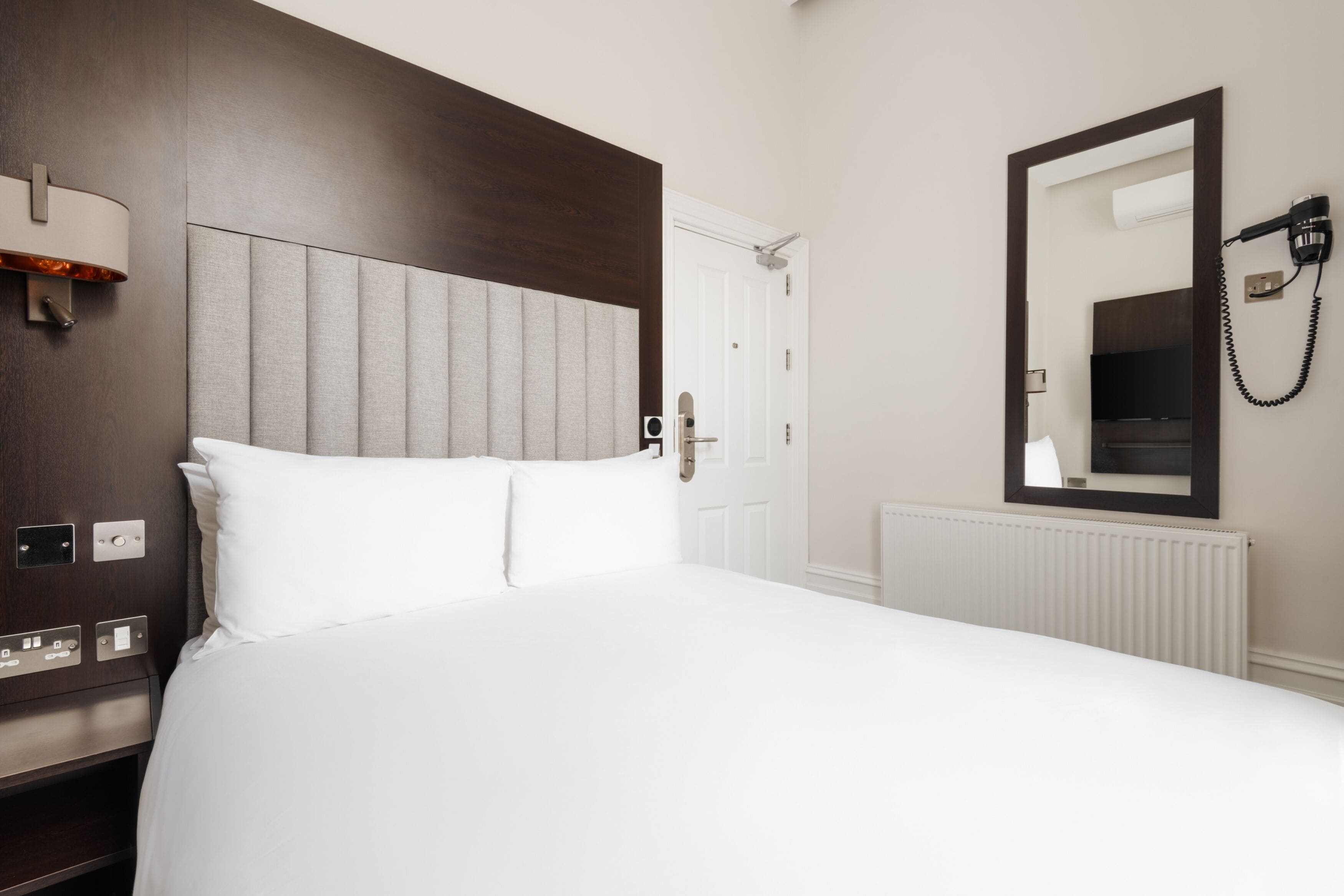A hotel room with a large bed, a headboard, a nightstand, a lamp, a mirror, and a hairdryer on the wall.