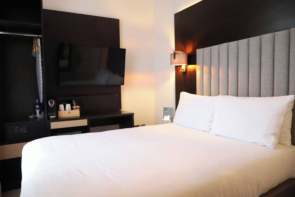 A hotel room with a large bed, a flat-screen TV mounted on the wall, a bedside table with a lamp, and a dark-colored headboard.