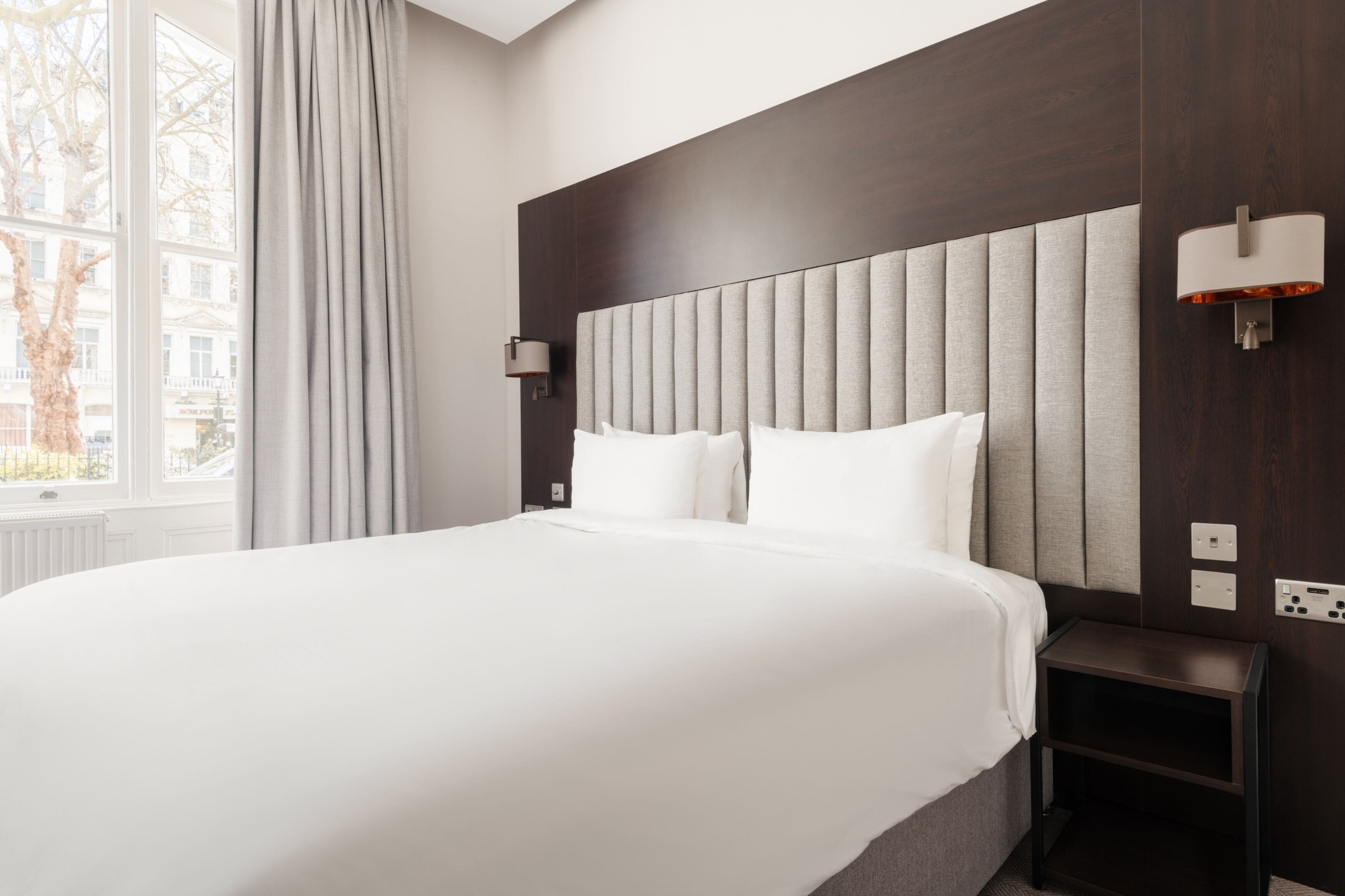 A hotel room with a large bed, a nightstand, a wall-mounted lamp, and a window with curtains.