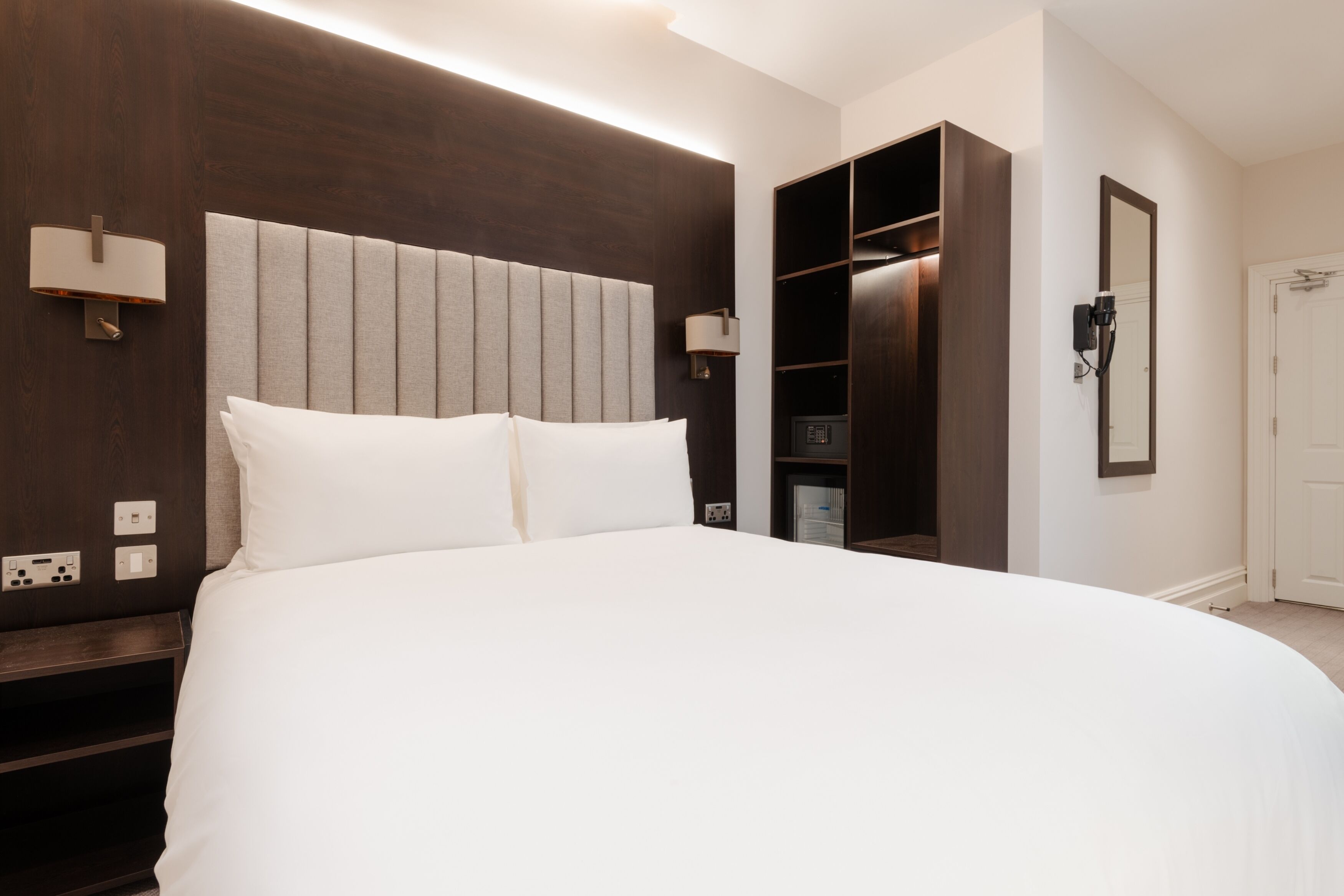 Une chambre d’hôtel moderne avec un grand lit, une armoire intégrée, des lampes de chevet et un miroir.