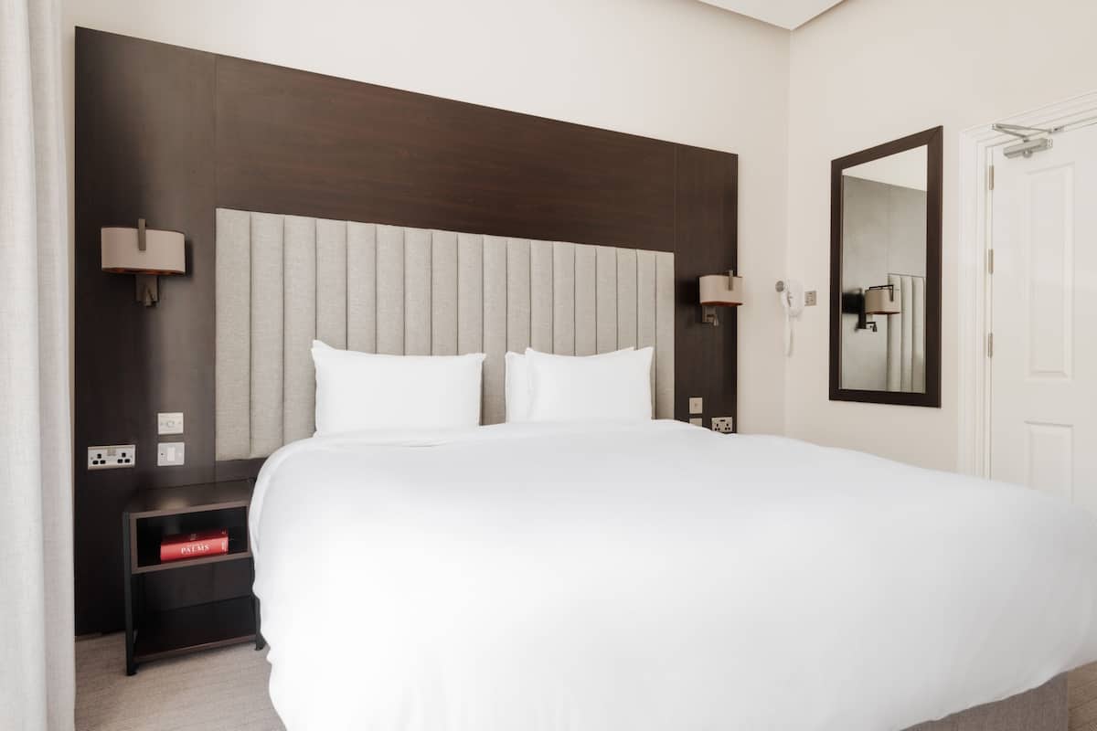 A hotel room with a large bed, a dark wood headboard, bedside lamps, a nightstand with a red book, and a mirror on the wall.