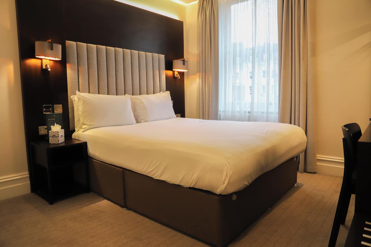 A modern hotel room with a large bed, two bedside lamps, a nightstand with a tissue box, and a view of the city through the window.