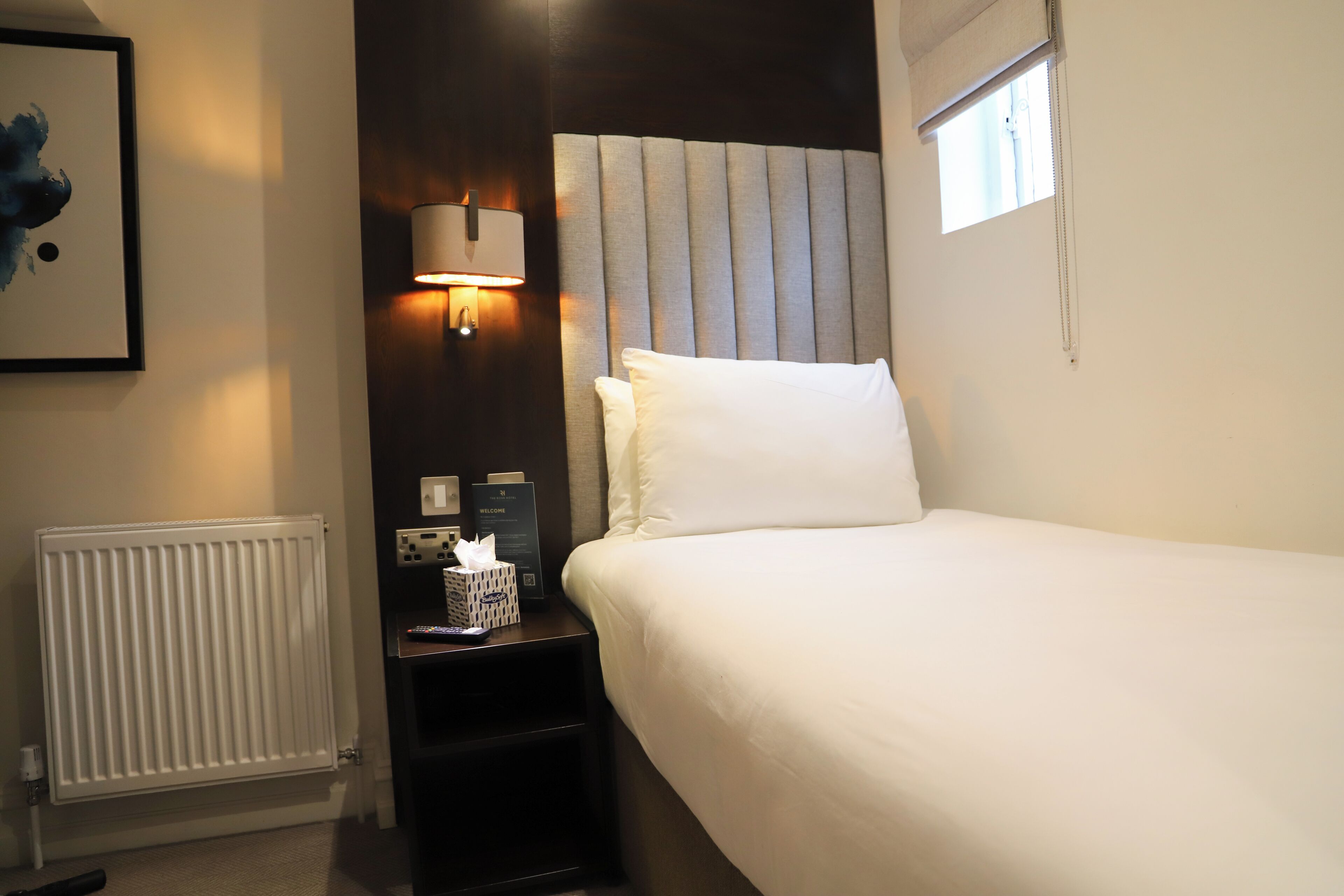 A hotel room with a large bed, a bedside table with a lamp, a radiator, and a framed picture on the wall.