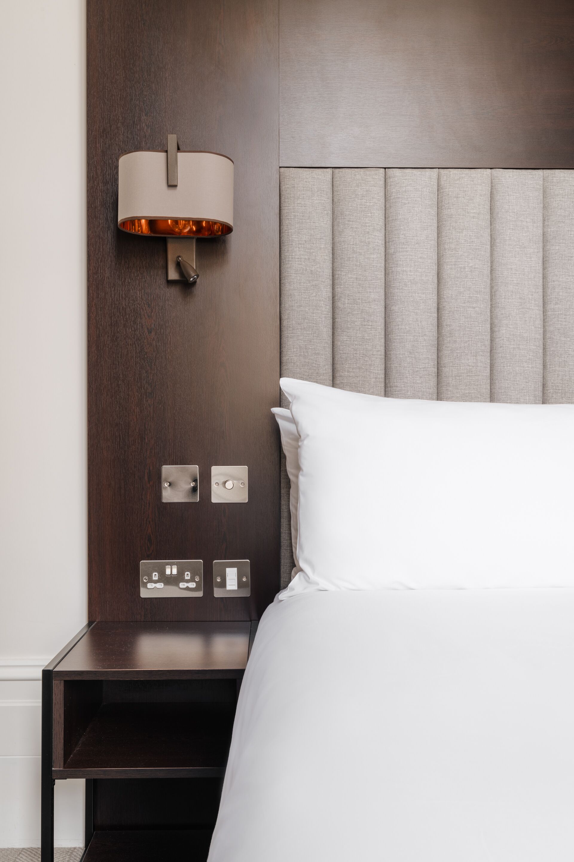 Habitación de hotel con cama, mesita de noche, lámpara de pared y panel de control.