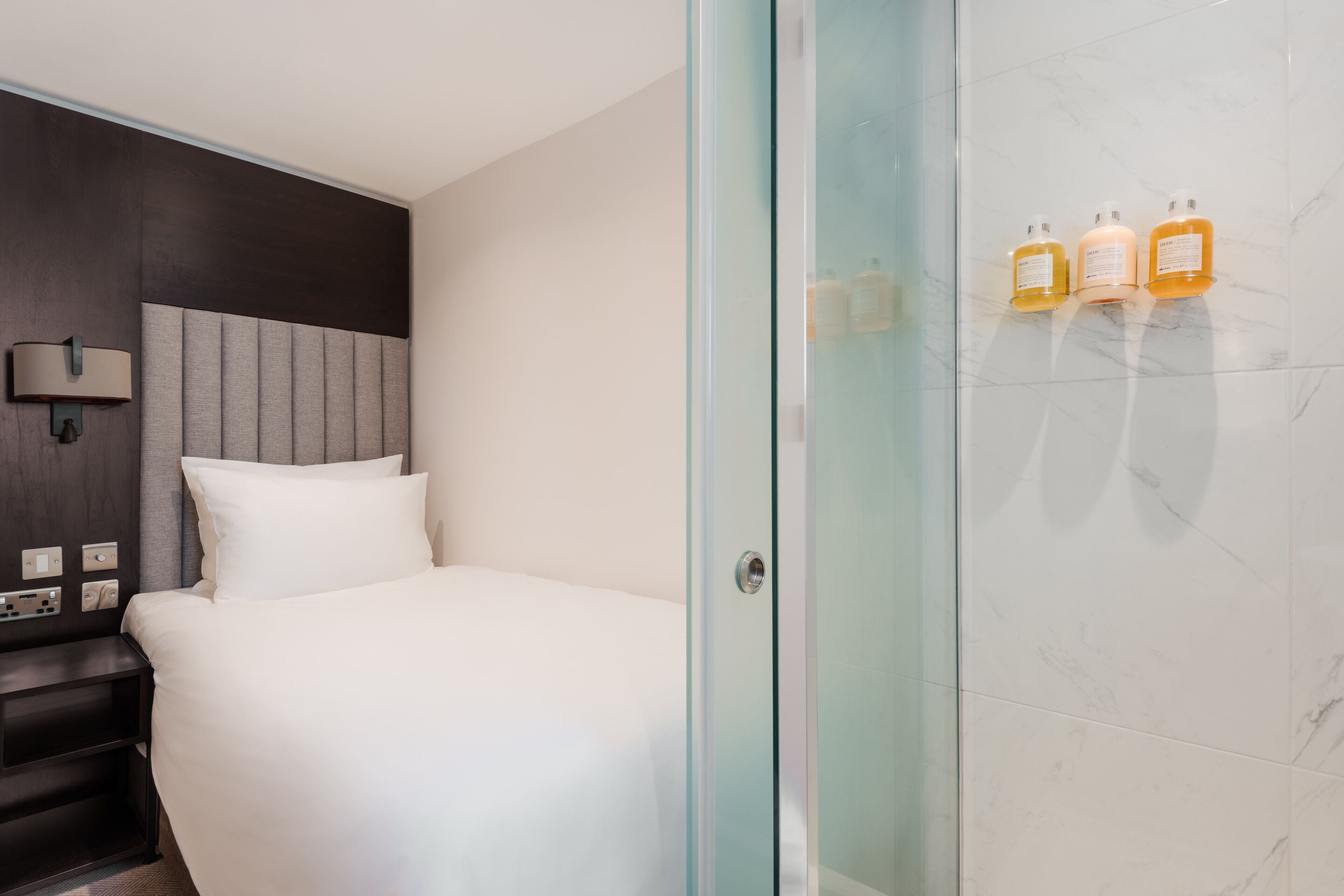 A hotel room with a bed, a nightstand, a wall-mounted lamp, and a glass shower enclosure with toiletries.