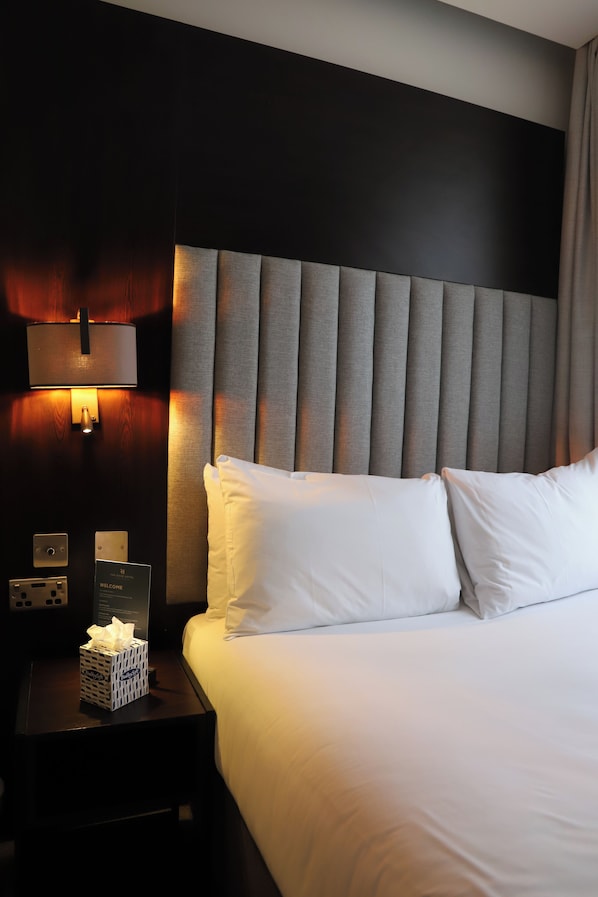 Ein Hotelzimmer mit einem großen Bett, einem Nachttisch mit Taschentuchbox und einer Wandlampe. Ein Schild mit der Aufschrift 'WELCOME'.