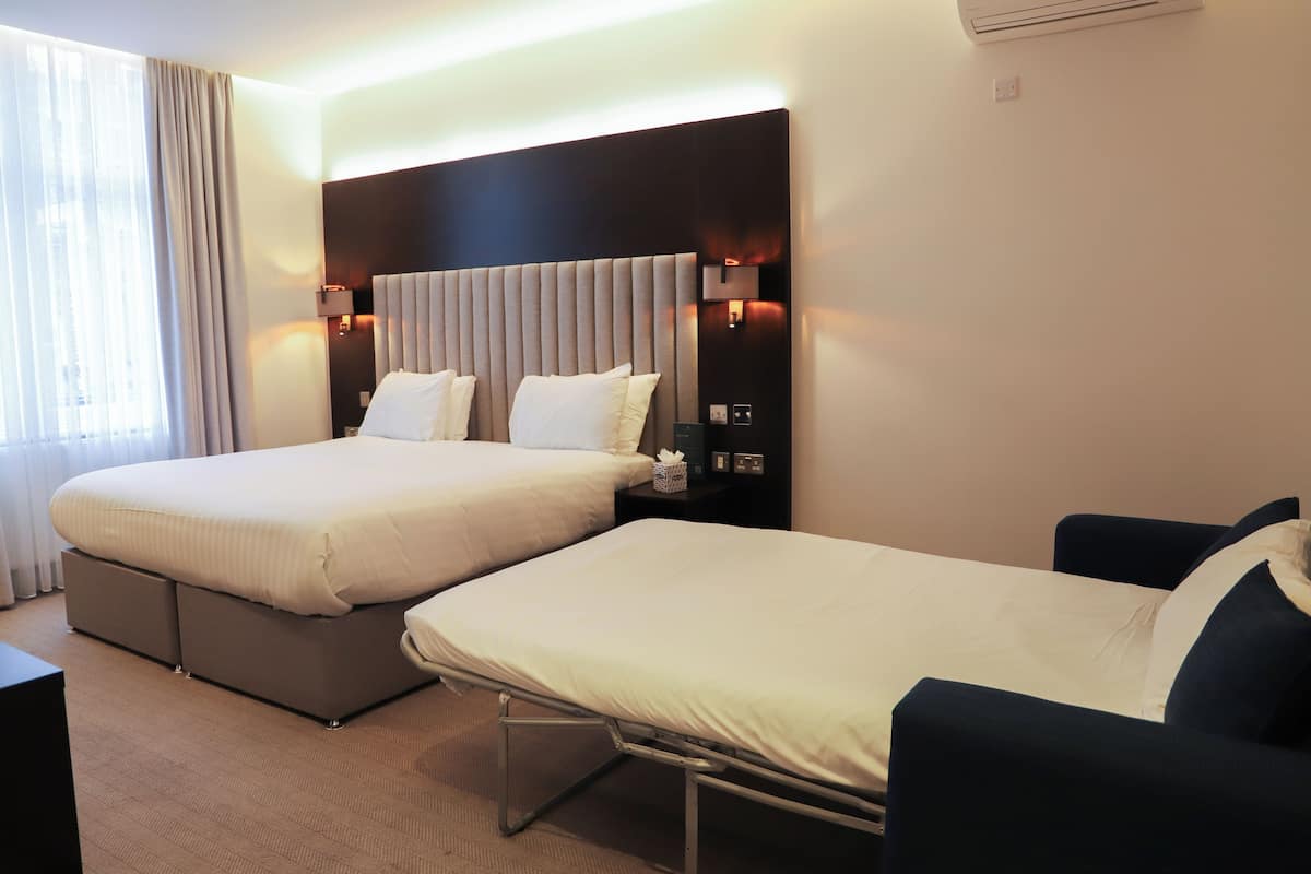 A hotel room with two beds, a dark headboard, bedside lamps, and a large window with curtains.