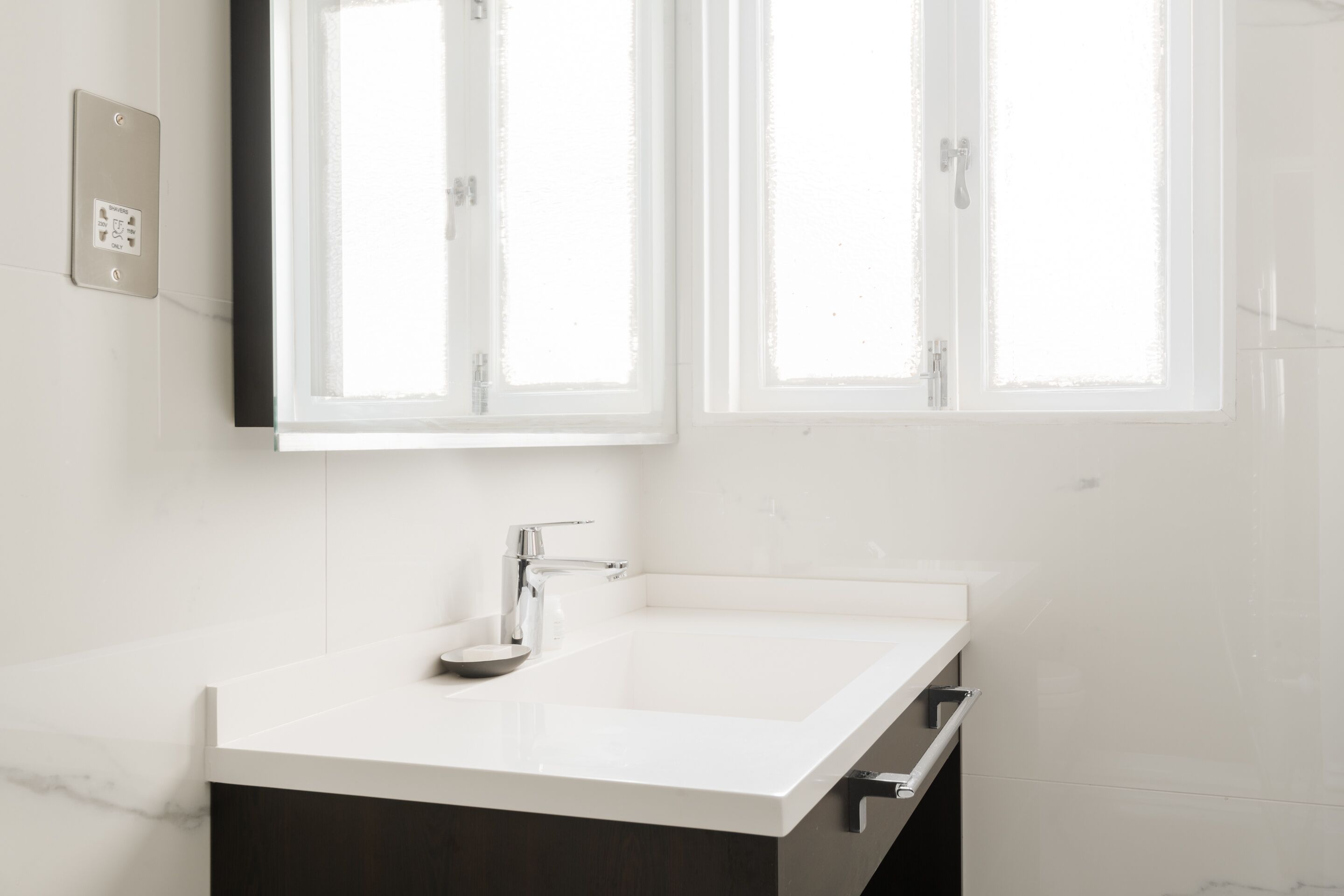 Une salle de bain moderne avec un lavabo blanc, des meubles de rangement aux couleurs foncées et une fenêtre située au-dessus du lavabo.