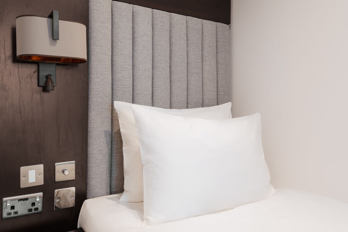 A hotel room with a bed, a nightstand, a wall-mounted lamp, and a control panel.