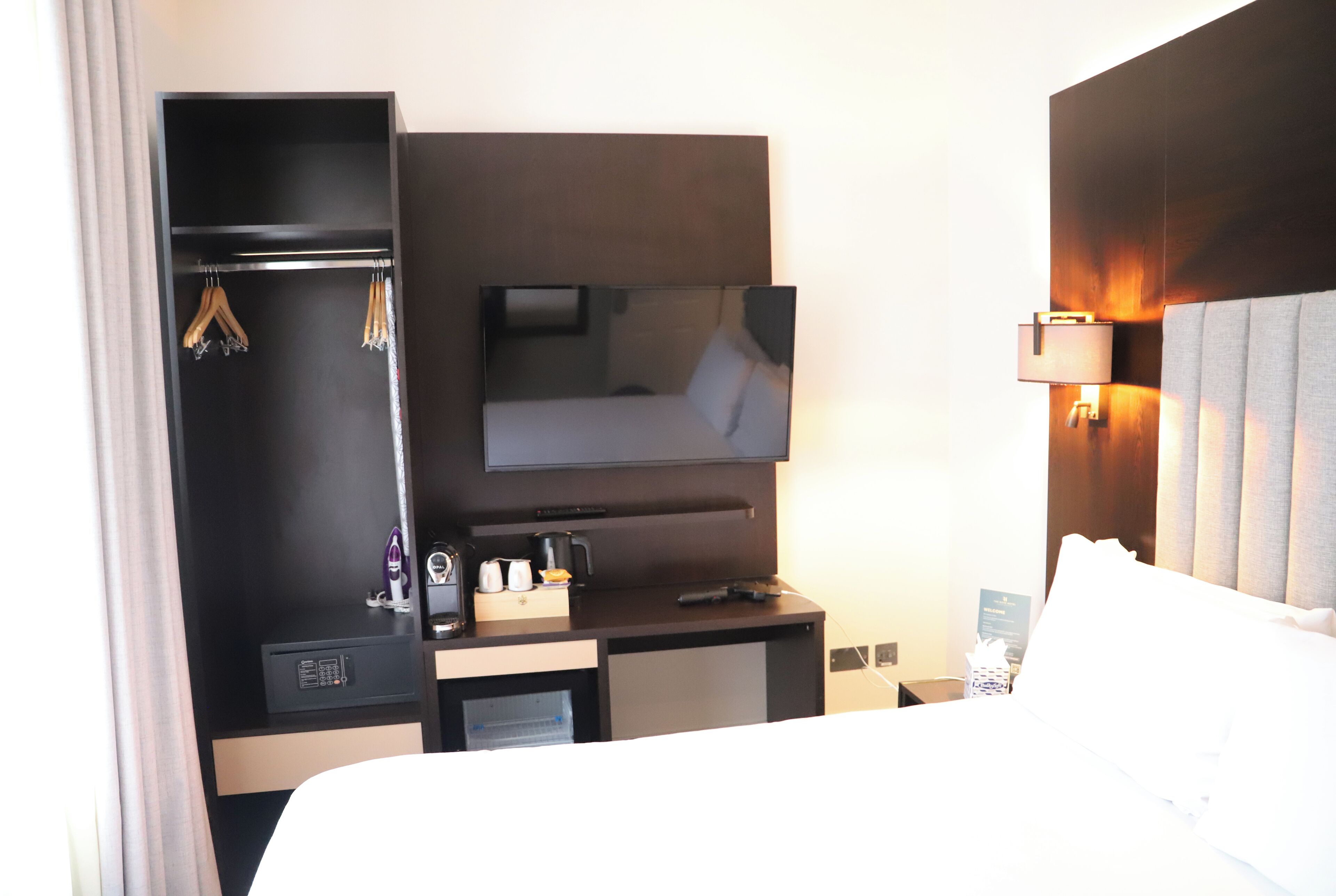 Une chambre d’hôtel comprenant un lit, un téléviseur fixé au mur, un bureau avec une machine à café et une armoire avec des vêtements suspendus.