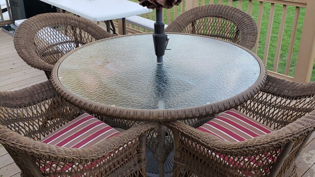 Une table ronde en verre, entourée de chaises en osier et agrémentée de coussins à rayures, est placée sur une terrasse extérieure équipée d’un barbecue et d’un parasol.