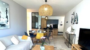 A modern living room with a sofa, coffee table, and TV. There is a dining area with chairs and a table, and a kitchen in the background. - SEAFRONT APARTMENT VAL ANDRE ALL INCLUSIVE FOR 5 PEOPLE (Pléneuf-Val-André)