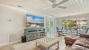 A spacious living room with a large TV, a wooden ceiling fan, a beige ottoman, and a leather couch.