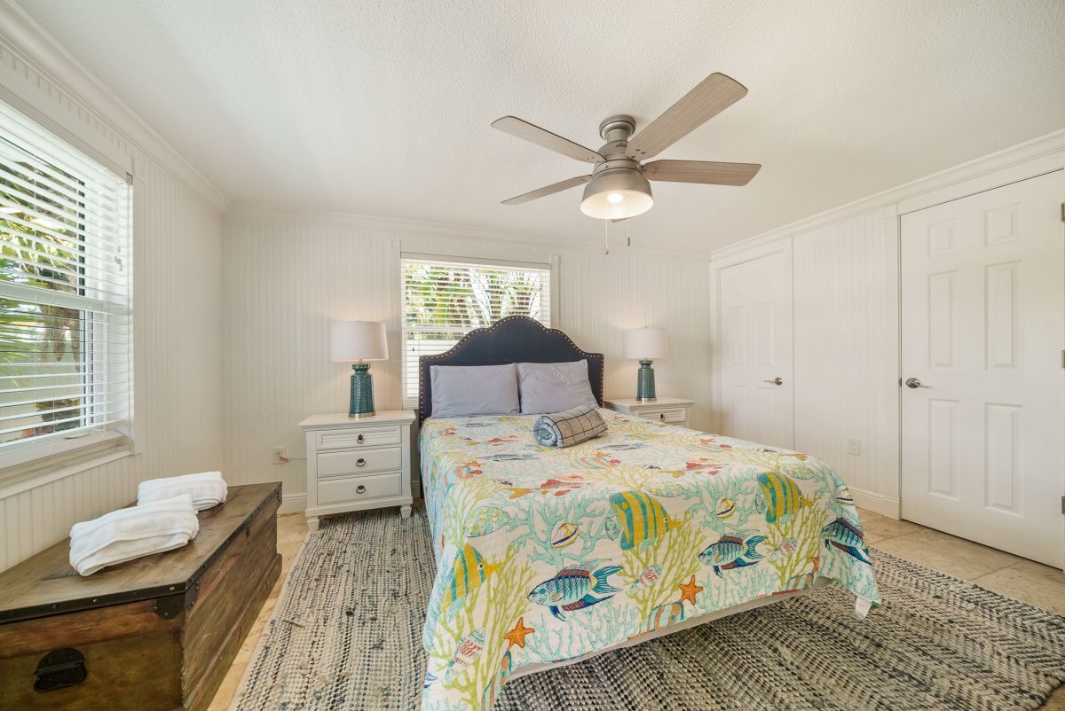 A bedroom with a bed, a ceiling fan, a window with blinds, a nightstand with a lamp, and a door.