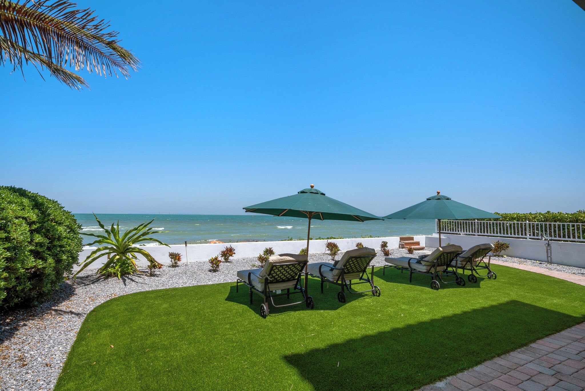 A patio with four lawn chairs and two umbrellas, overlooking a beach.