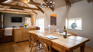 A spacious kitchen with wooden beams, a large wooden table, and a cozy seating area with a sofa and chairs. - Rural Barn Conversion (Truro)