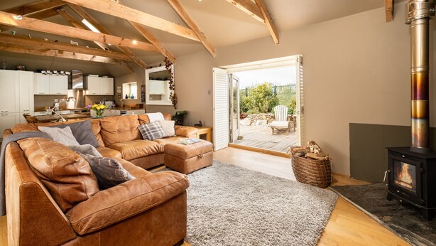 A cozy living room with a brown leather sofa, a fireplace, and a view of the outdoor patio. - Rural Barn Conversion (Truro)