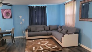 Ein Wohnzimmer mit einem Ecksofa, einem Esstisch mit Stühlen, einem Teppich und einem Deckenventilator.