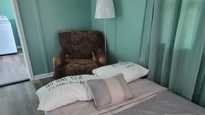 Ein Bett mit zwei Kissen, eines mit Schriftzug und das andere mit Streifenmuster. Daneben ein Stuhl mit Blumenmuster und eine weiße Lampe.