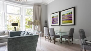 Uma sala de estar moderna com um sofá cinza, uma mesa de centro de vidro e duas poltronas cinzas. Há duas pinturas emolduradas na parede, um candelabro e uma janela com cortinas.